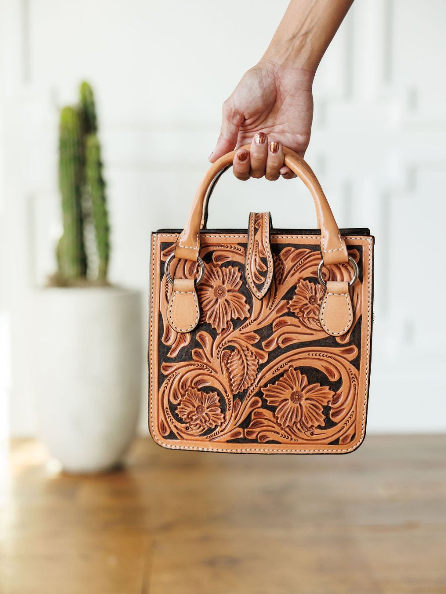 A hand holds The Campitos, a small, rectangular floral-embossed leather handbag by Savannah Sevens western life{&}style. In the blurred background, a potted cactus rests on a wooden floor.