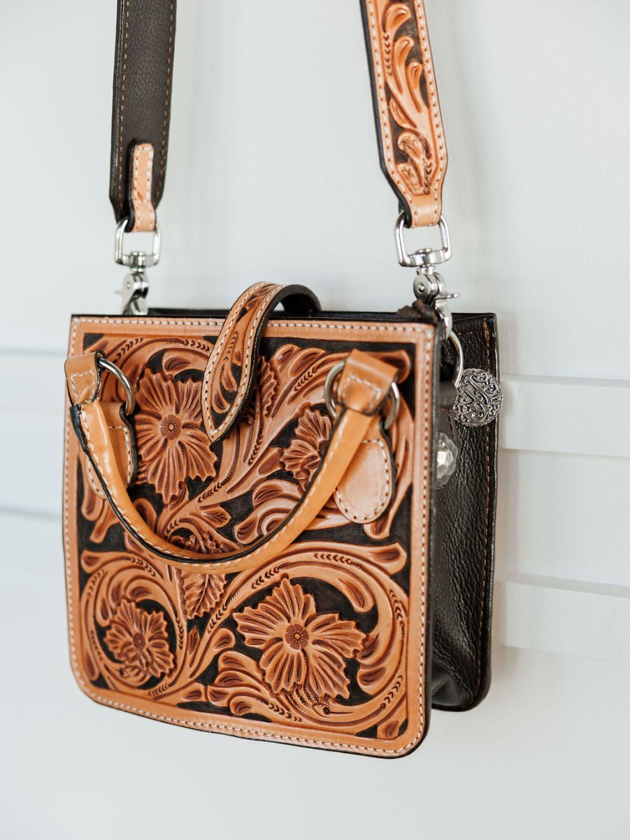 The Campitos by Savannah Sevens western life{&}style is a floral tooled leather handbag in tan and dark brown with flower and leaf patterns, a shoulder strap, and two handles. It hangs on a white wall for display. By Double J.