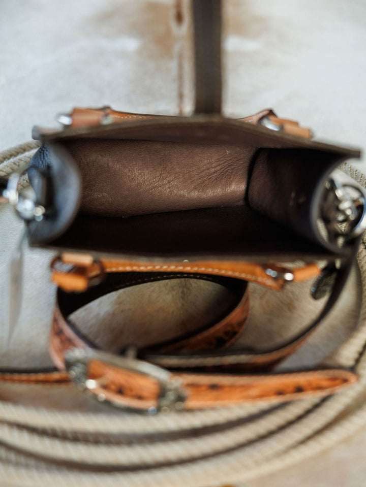 A close-up of The Campitos hand tooled leather handbag by Savannah Sevens western life{&}style, featuring intricate stitching and metal buckle accents, displays its empty interior on coiled rope atop a light surface. By Double J. 