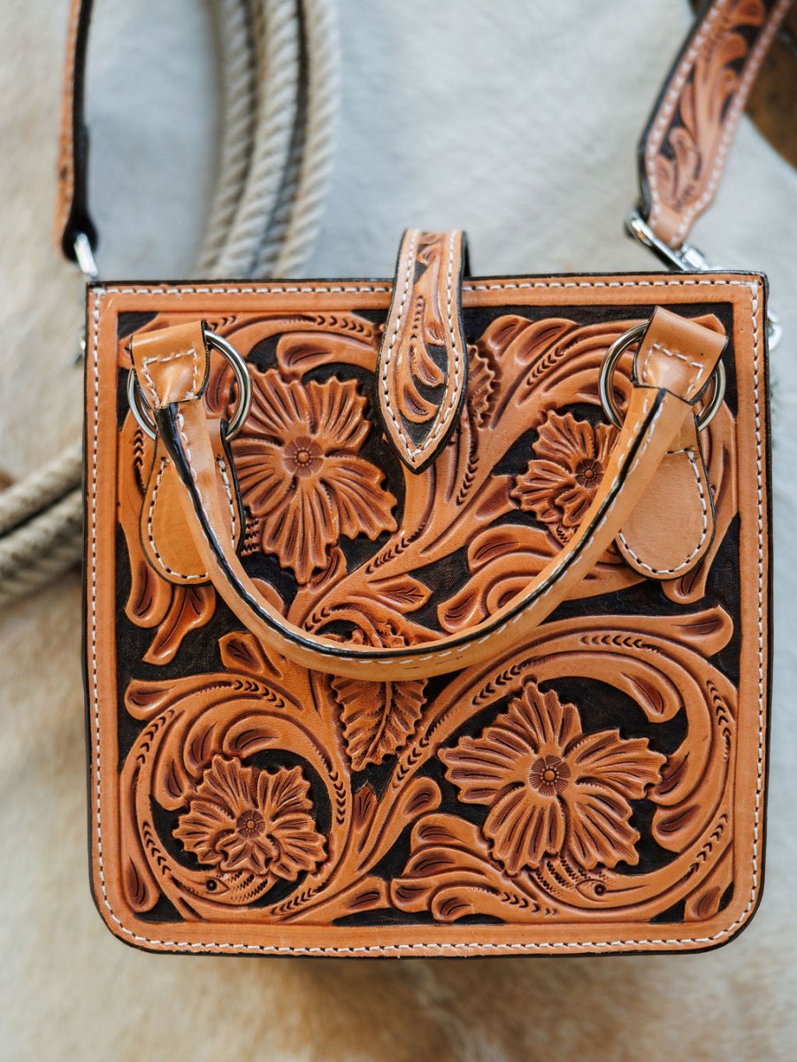 The Campitos by Savannah Sevens western life{&}style is a square, hand-tooled leather handbag with embossed floral and swirling designs, a short handle, snap closure, plus a coiled rope and light surface in the background. By Double J. 