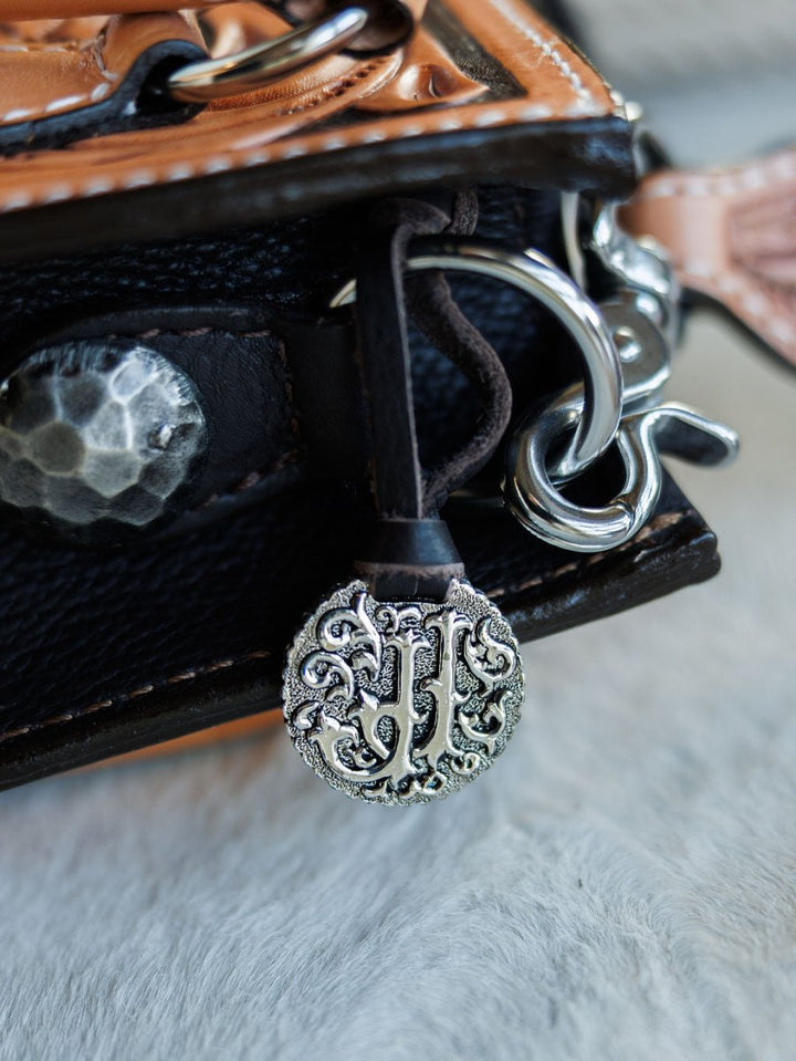 A close-up of a textured metal charm with ornate designs and “JH” initials on The Campitos handbag by Savannah Sevens western life{&}style, featuring hand tooled black and brown leather with silver hardware. By Double J. 