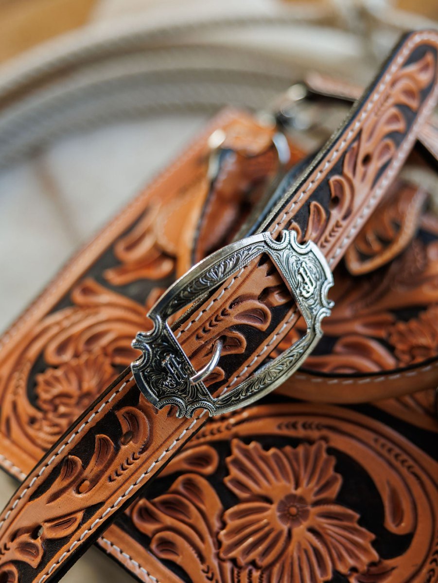 Close-up of The Campitos by Savannah Sevens western life{&}style: intricately tooled brown leather straps with ornate silver buckles and detailed floral scrollwork, perfect for a hand-tooled leather handbag. Blurred coiled rope in the background. By Double J. 