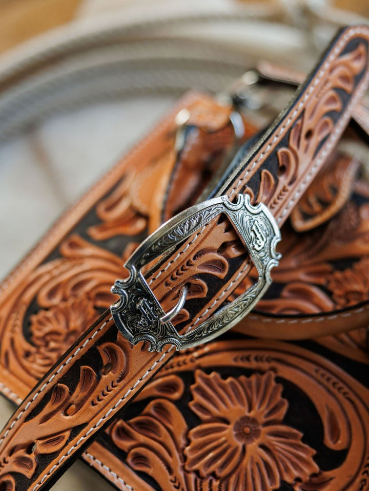 Close-up of The Campitos by Savannah Sevens western life{&}style: intricately tooled brown leather straps with ornate silver buckles and detailed floral scrollwork, perfect for a hand-tooled leather handbag. Blurred coiled rope in the background. By Double J. 