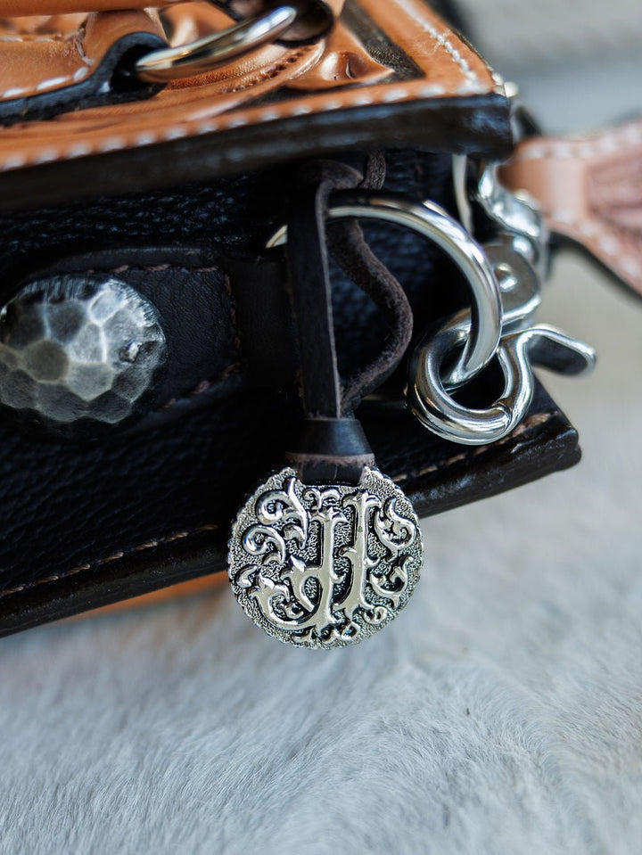 A close-up of a textured metal charm with ornate designs and “JH” initials on The Campitos handbag by Savannah Sevens western life{&}style, featuring hand tooled black and brown leather with silver hardware. By Double J. 