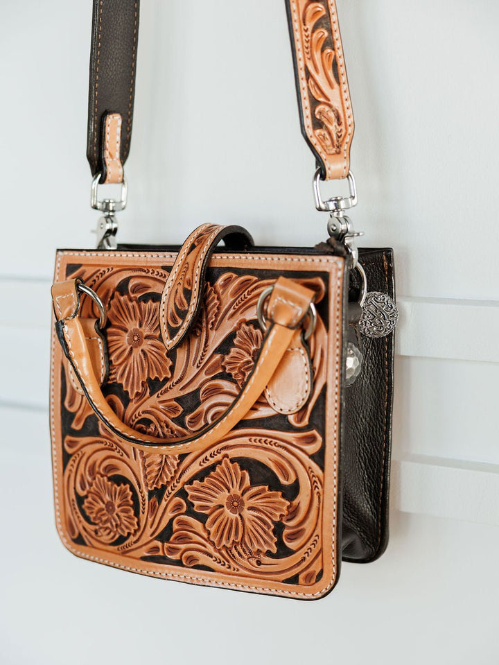 The Campitos by Savannah Sevens western life{&}style is a floral tooled leather handbag in tan and dark brown with flower and leaf patterns, a shoulder strap, and two handles. It hangs on a white wall for display. By Double J.