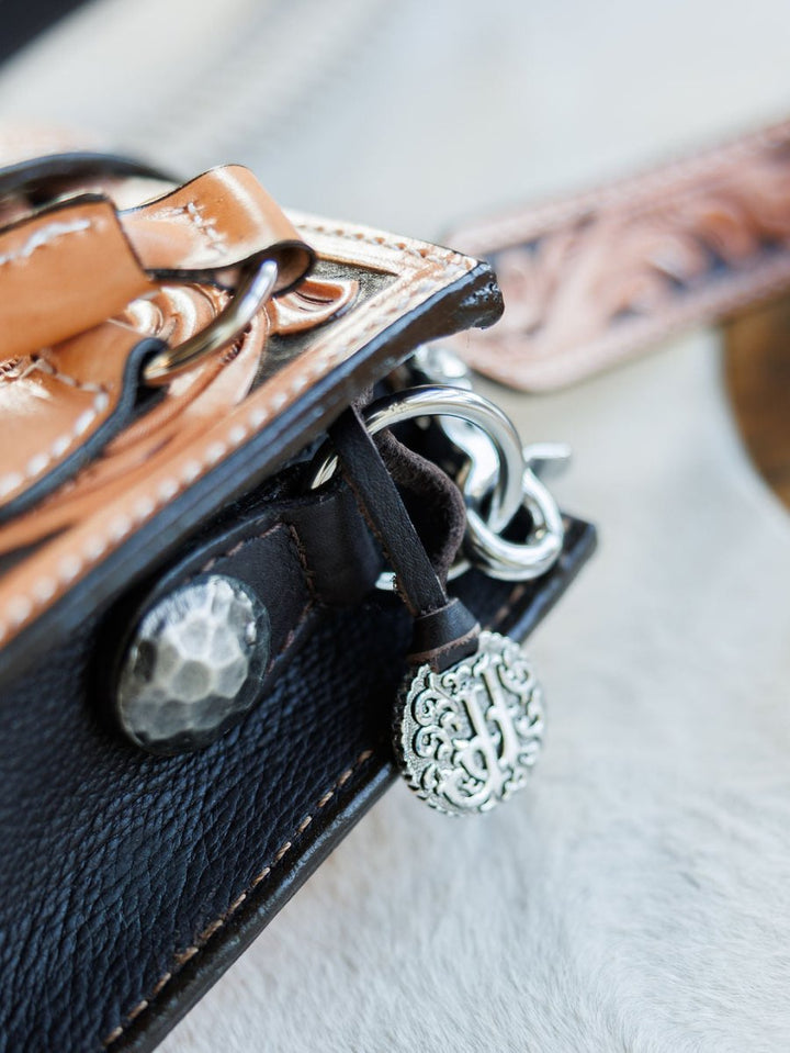 Close-up of The Campitos floral leather handbag by Savannah Sevens western life{&}style, with a decorative silver button, metal clasp, and round tree-design charm. Detailed leatherwork strap. Bag rests on a soft, light-colored surface. By Double J. 