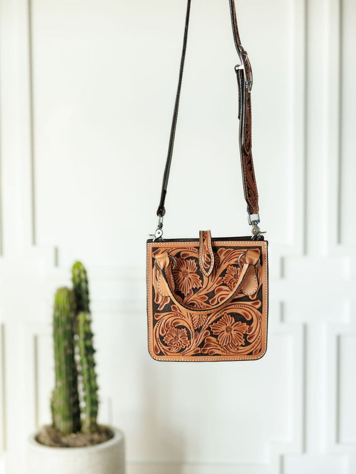 The Campitos floral leather handbag by Savannah Sevens western life{&}style, featuring detailed embossing, hangs before a white wall with a potted cactus seen in the left background.