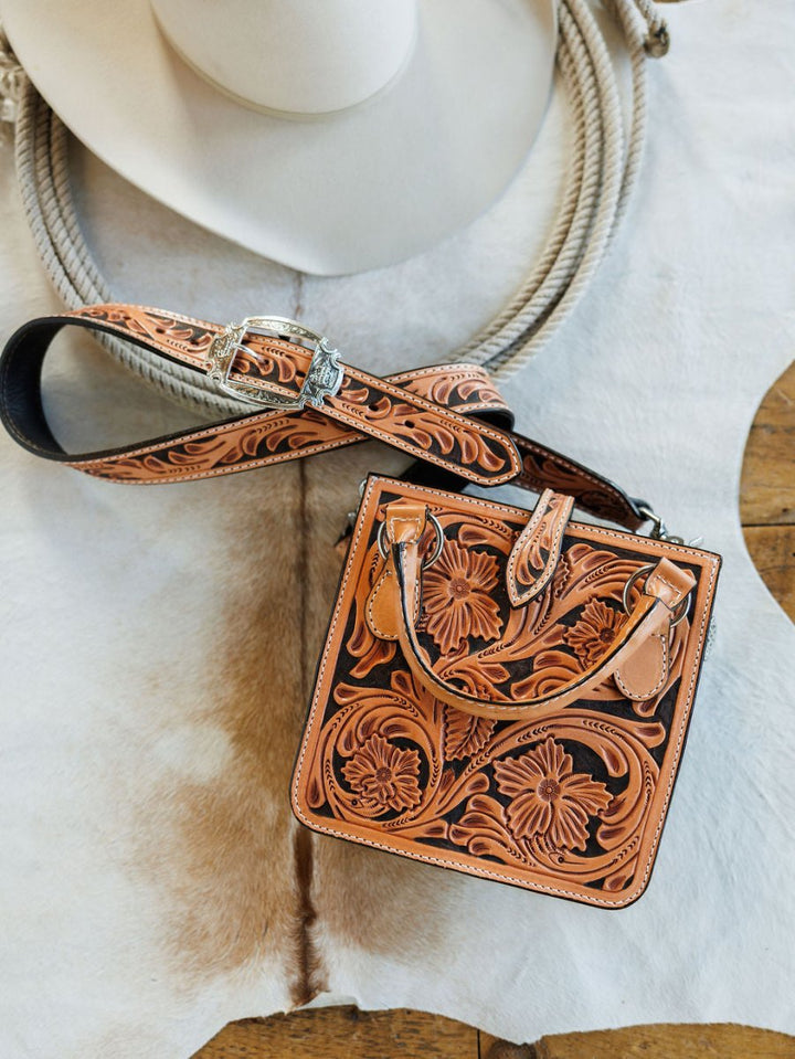 The Campitos leather handbag from Savannah Sevens western life{&}style, featuring floral designs, rests on a cowhide rug next to a white cowboy hat.