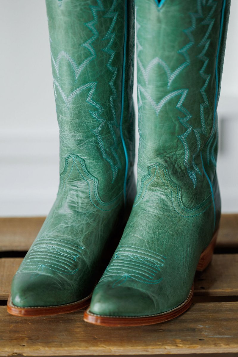 A close-up of The Carico by Savannah Sevens western life{&}style: a green turquoise leather western boot with blue stitching, pointed toes, a tall patterned shaft, shown on a wooden surface.