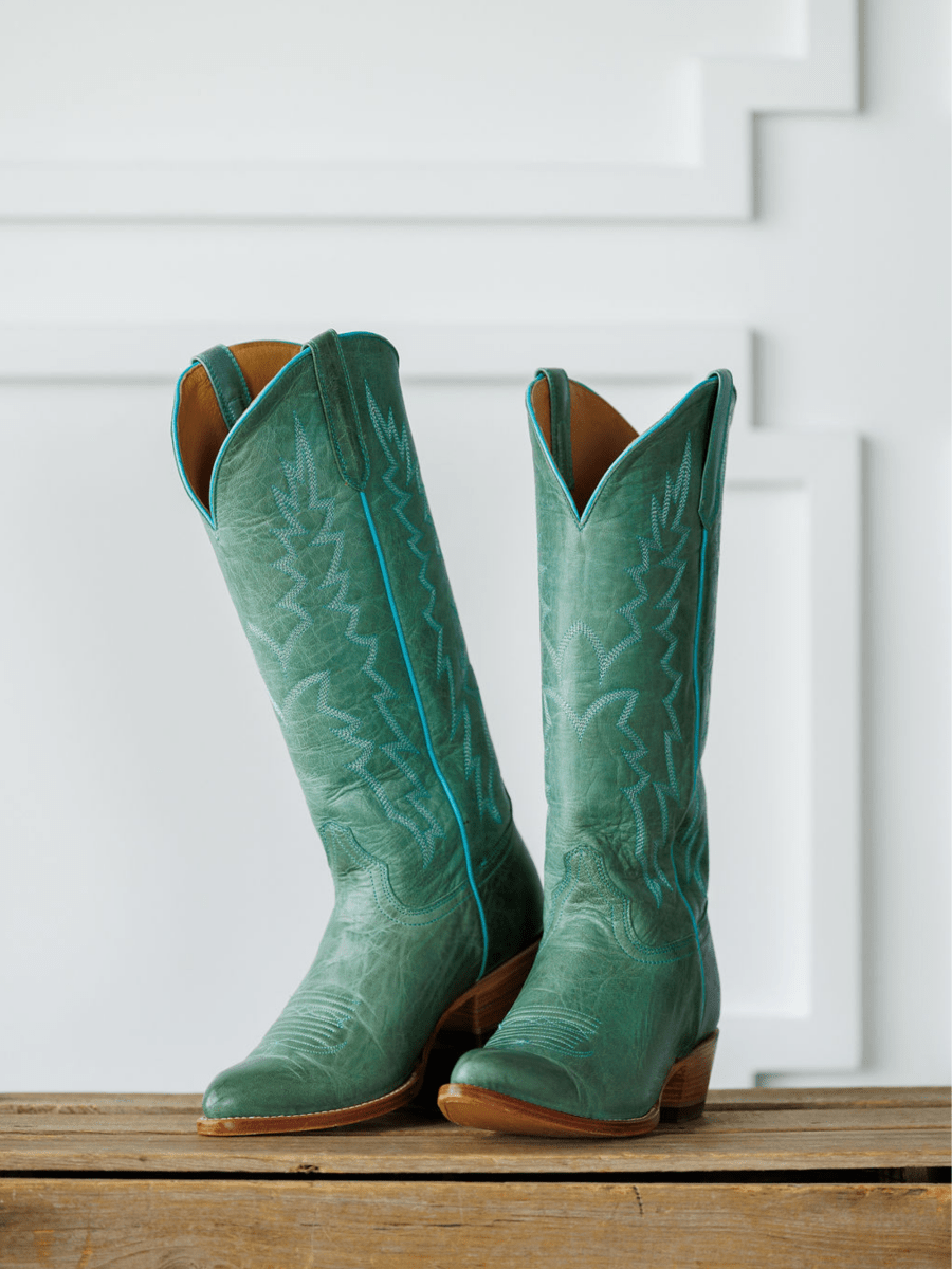 The Carico western boots by Savannah Sevens western life{&}style, featuring tall green turquoise leather with subtle embroidery and an almond toe, stand side by side on a wooden surface against a white paneled background. Made by Black Star. 