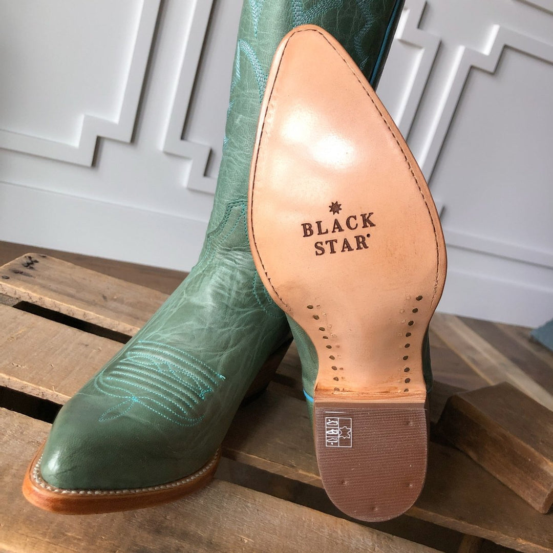 A close-up of The Carico, a green turquoise leather western boot with an almond toe from Savannah Sevens western life{&}style, rests on a wooden pallet. The tan sole shows the Black Star brand, with white wall paneling in the background. Made by Black Star. 
