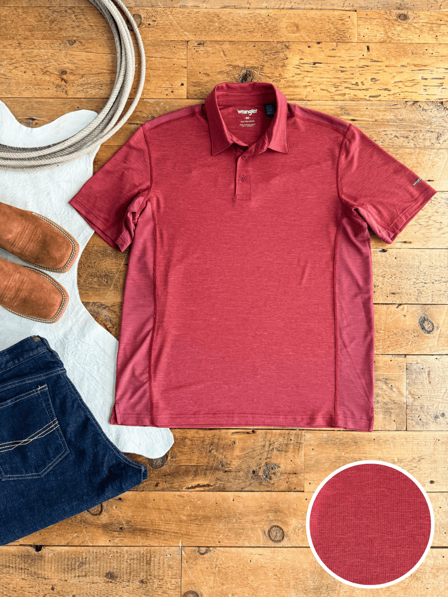 A red "The Carlyle" men's polo by Savannah Sevens western life{&}style, made from performance fabric, lies flat on a wooden floor with brown boots, dark blue jeans, and a white saddle pad. A close-up highlights the shirt’s texture.