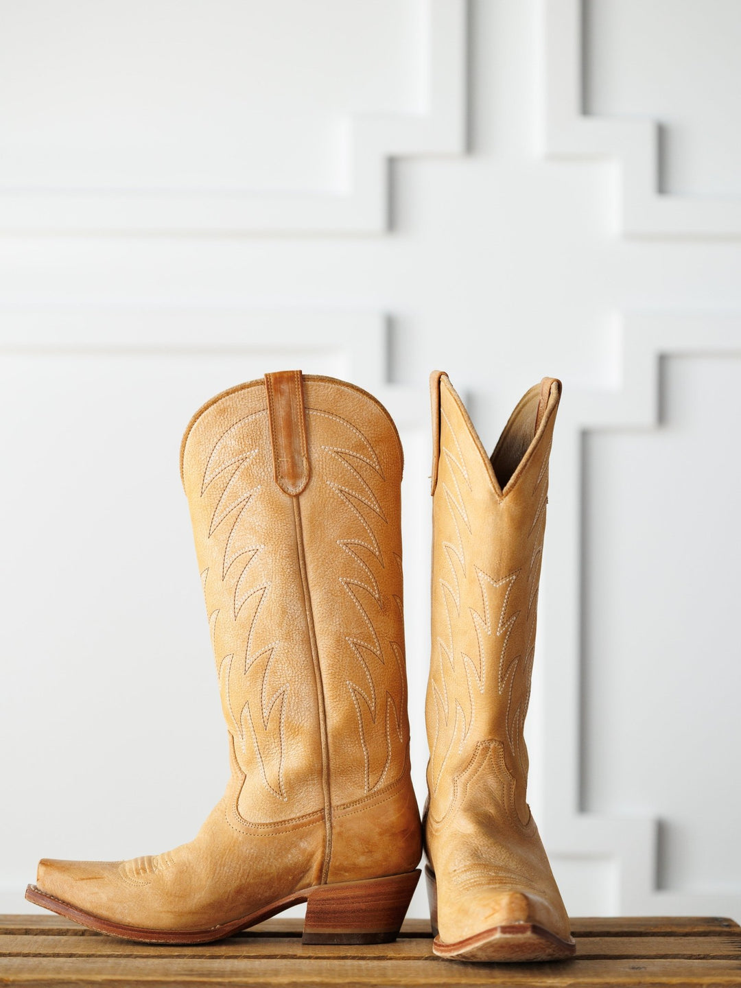 The Carmencita western boots by Savannah Sevens western life{&}style, in saddle tan leather with decorative stitching—one upright, one on its side—are displayed on a wooden surface against a white patterned background.