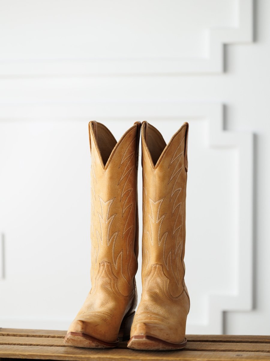 The Carmencita by Savannah Sevens western life{&}style: saddle tan leather western boots with decorative stitching stand upright on a wooden surface against a white, textured background.