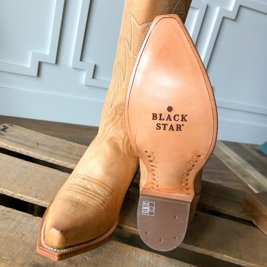 The Carmencita boot by Savannah Sevens western life{&}style, in saddle tan leather with a snip toe and decorative stitching, rests on a wooden pallet sole-up to reveal the BLACK STAR logo stamped on its smooth leather.
