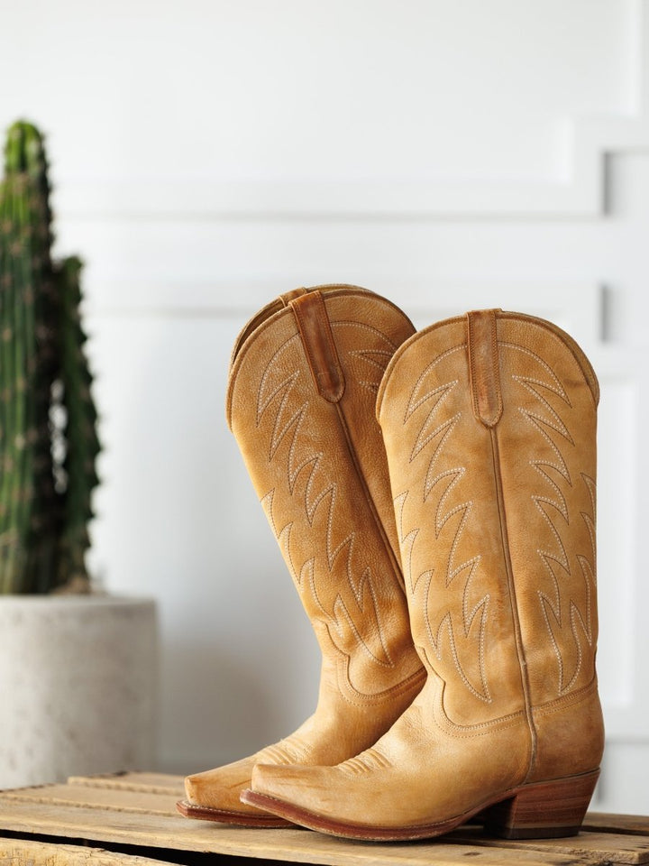 The Carmencita western boots by Savannah Sevens western life{&}style in saddle tan leather, featuring decorative stitching, rest on a wooden surface with a tall green cactus in a white pot behind them. Boots made by Blackstar. 