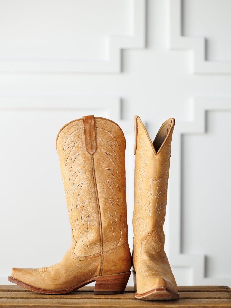 The Carmencita western boots by Savannah Sevens western life{&}style, in saddle tan leather with decorative stitching—one upright, one on its side—are displayed on a wooden surface against a white patterned background.