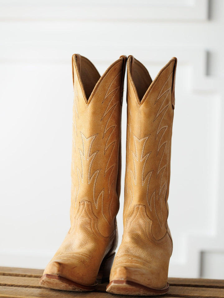 The Carmencita boots by Savannah Sevens western life{&}style, crafted in saddle tan leather with decorative stitching, stand side by side on a wooden surface against a white background. Boots made by Black Star. 