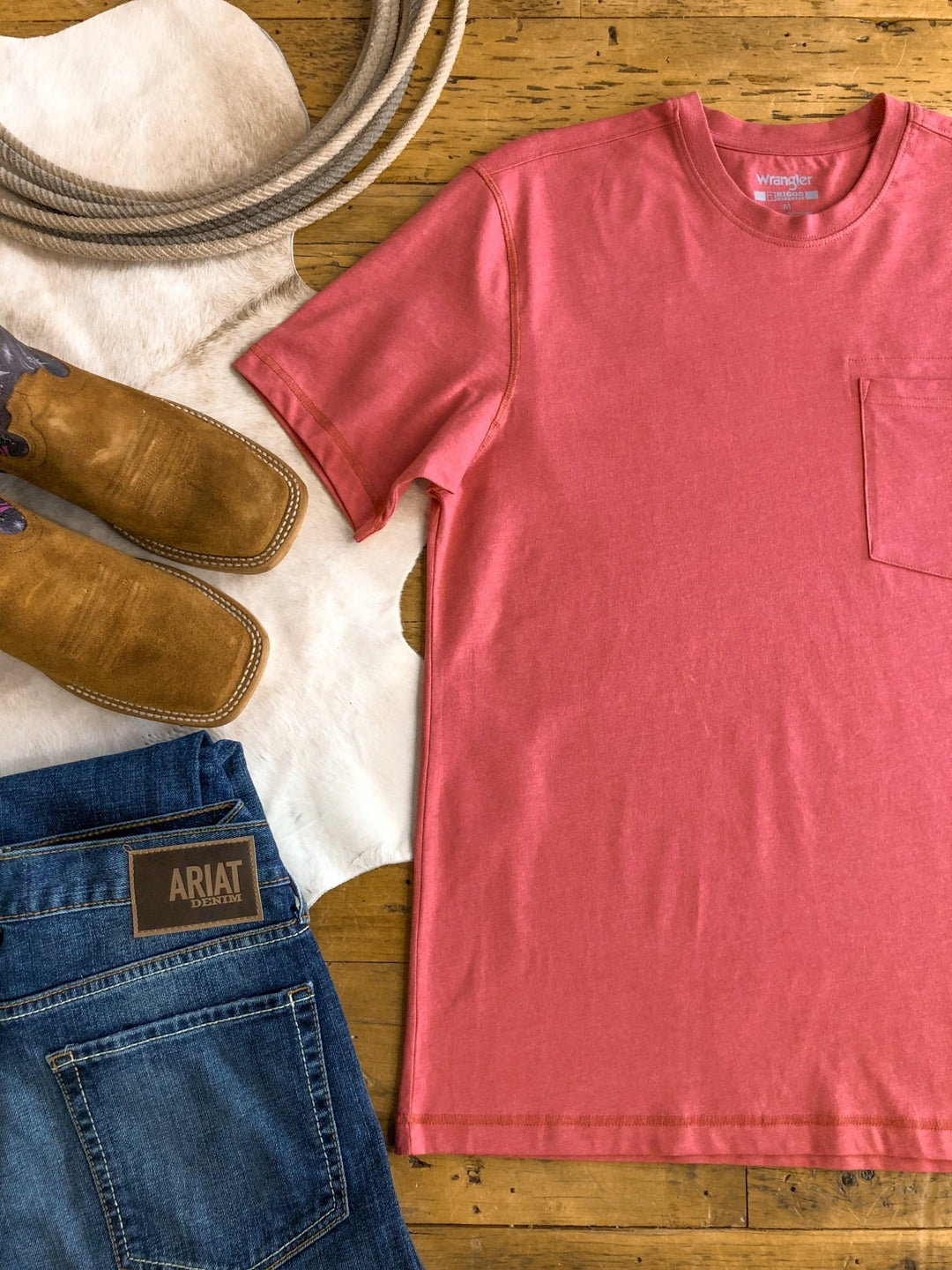 Flat lay featuring The Carver {Men’s} tee (XL) by Savannah Sevens western life{&}style, blue Ariat jeans, tan cowboy boots, a white cowhide, and a coiled rope arranged on a wooden floor.