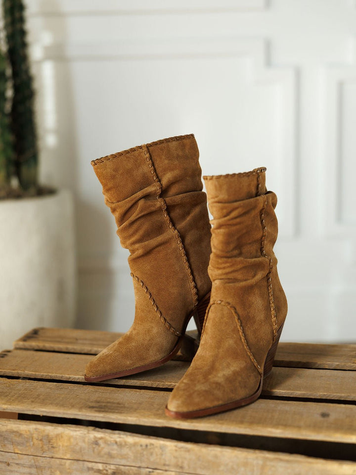 The Cerritos by Savannah Sevens western life{&}style—tan suede slouchy boots with pointed toes, decorative stitching, and a stacked heel—are displayed on a wooden pallet with a white wall and potted cactus in the background.