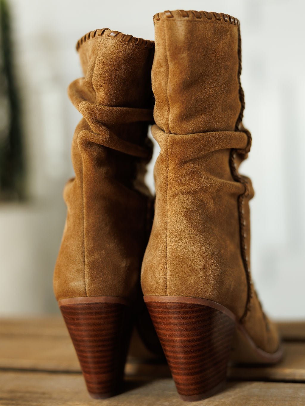 The Cerritos suede bootie by Savannah Sevens western life{&}style features a stacked heel, visible stitching, and a soft, slouchy texture, shown in a close-up on a wooden surface.