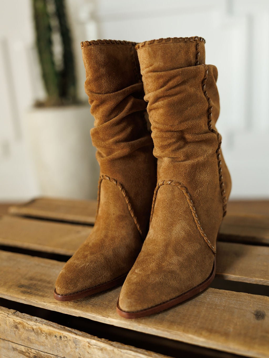 The Cerritos by Savannah Sevens western life{&}style are brown suede booties with stacked heels, a slouchy pointed toe, and visible stitching, shown on wood with a blurred potted cactus in the background.