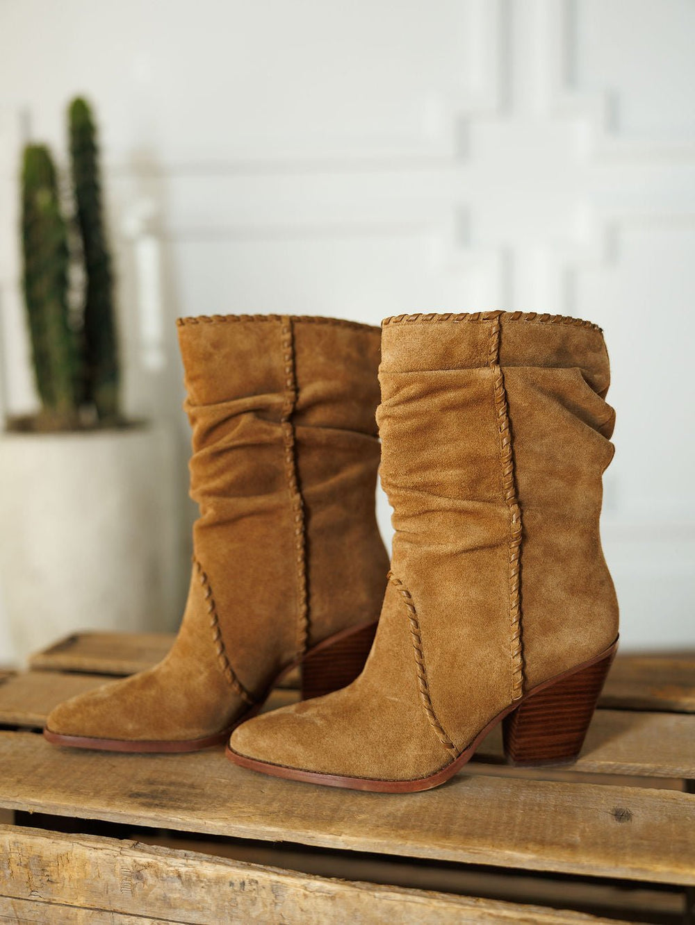 The Cerritos by Savannah Sevens western life{&}style: tan suede booties with slouchy shafts and stacked heels, shown on a wooden surface with a blurred potted cactus in the background.