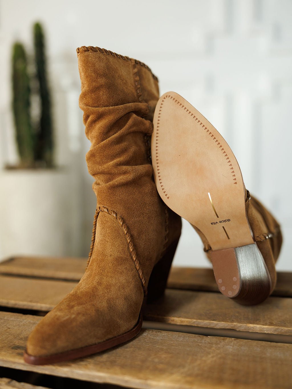 The Cerritos by Savannah Sevens western life{&}style: tan suede slouchy boots with stacked heels are shown on a wooden surface—one upright, the other revealing its sole. A blurred cactus is in the background.