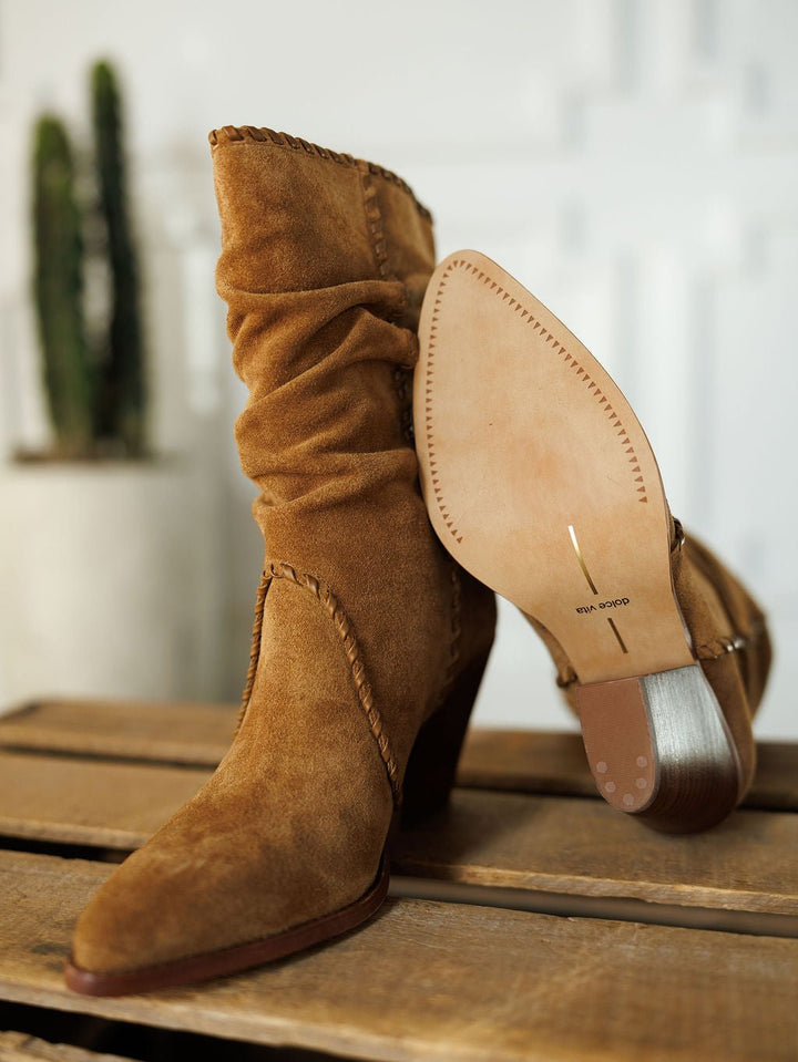 The Cerritos by Savannah Sevens western life{&}style: tan suede slouchy boots with stacked heels are shown on a wooden surface—one upright, the other revealing its sole. A blurred cactus is in the background.
