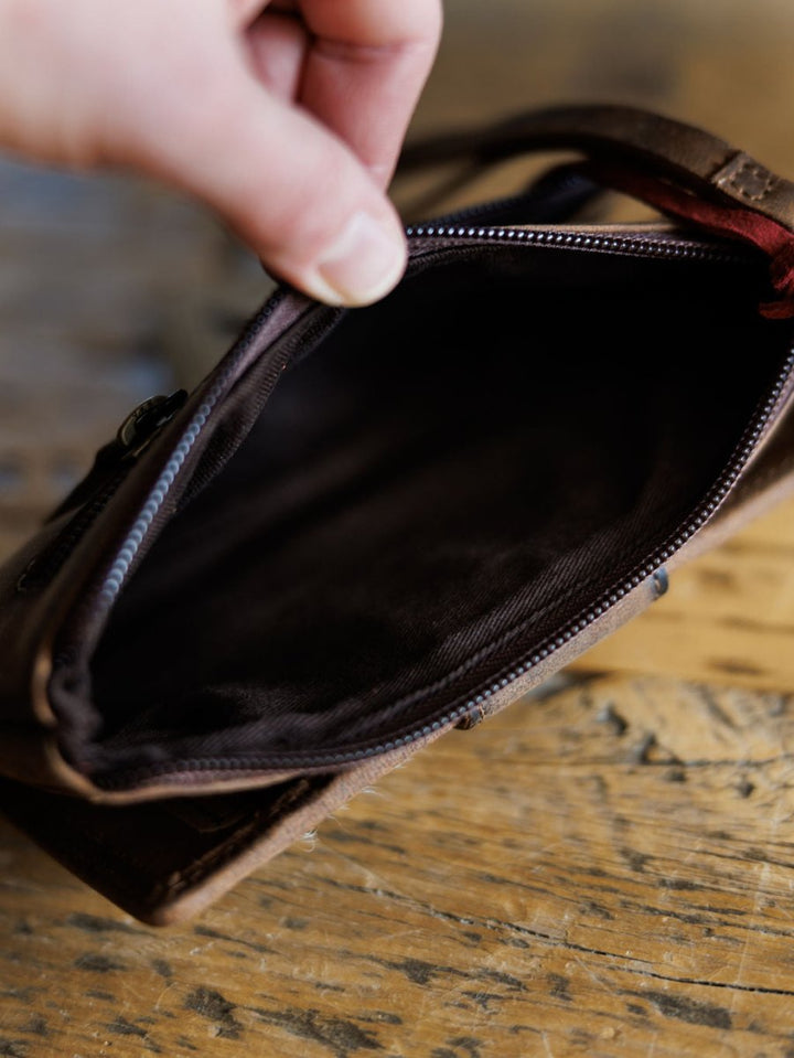 A hand unzips The Chisholm cowhide wristlet wallet by Savannah Sevens western life{&}style, showing its dark fabric interior against a wooden surface. By Sts. 