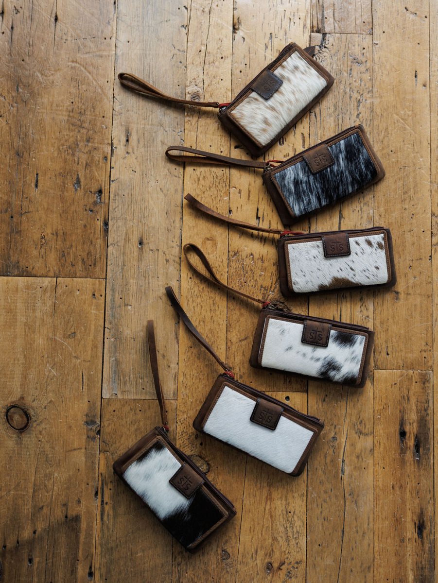Six “The Chisholm” wristlet wallets by Savannah Sevens western life{&}style, in black, white, and brown cowhide patterns, are arranged diagonally on a rustic wooden floor. By STS. 