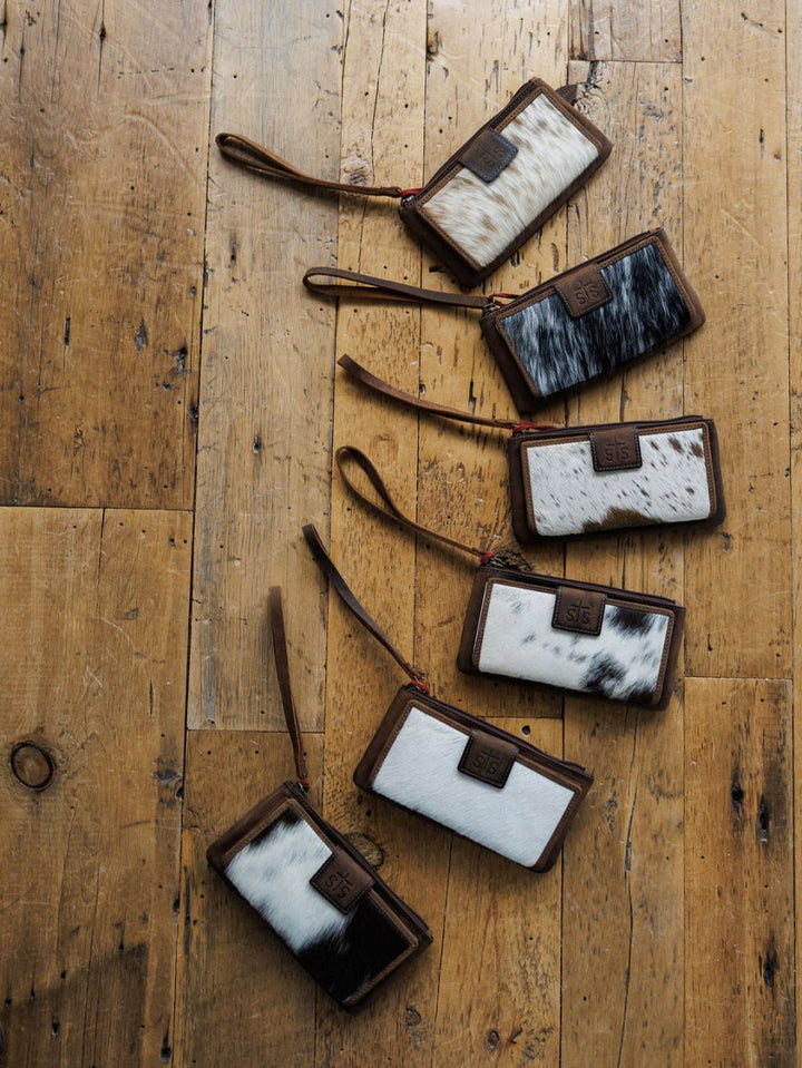 Six “The Chisholm” wristlet wallets by Savannah Sevens western life{&}style, in black, white, and brown cowhide patterns, are arranged diagonally on a rustic wooden floor. By STS. 