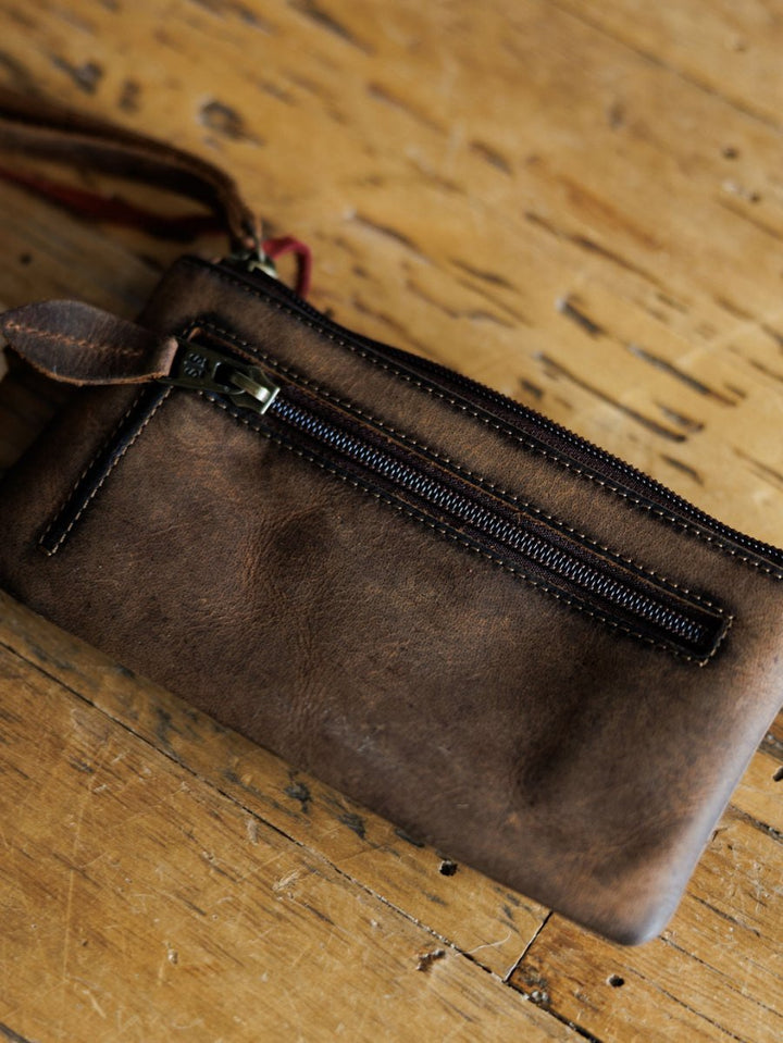 The Chisholm cowhide wristlet wallet by Savannah Sevens western life{&}style, featuring a zipper pocket and fabric loop, rests on a worn wooden surface.