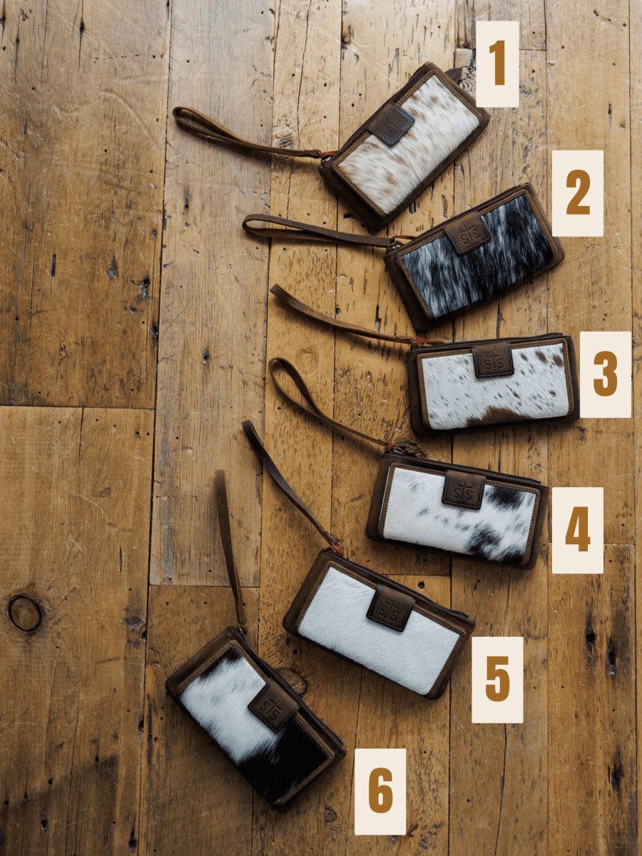 Six "The Chisholm" cowhide wristlet wallets by Savannah Sevens western life{&}style, each with brown leather trim and unique patterns, are arranged diagonally on a rustic wooden floor and labeled 1 to 6 with white numbers.