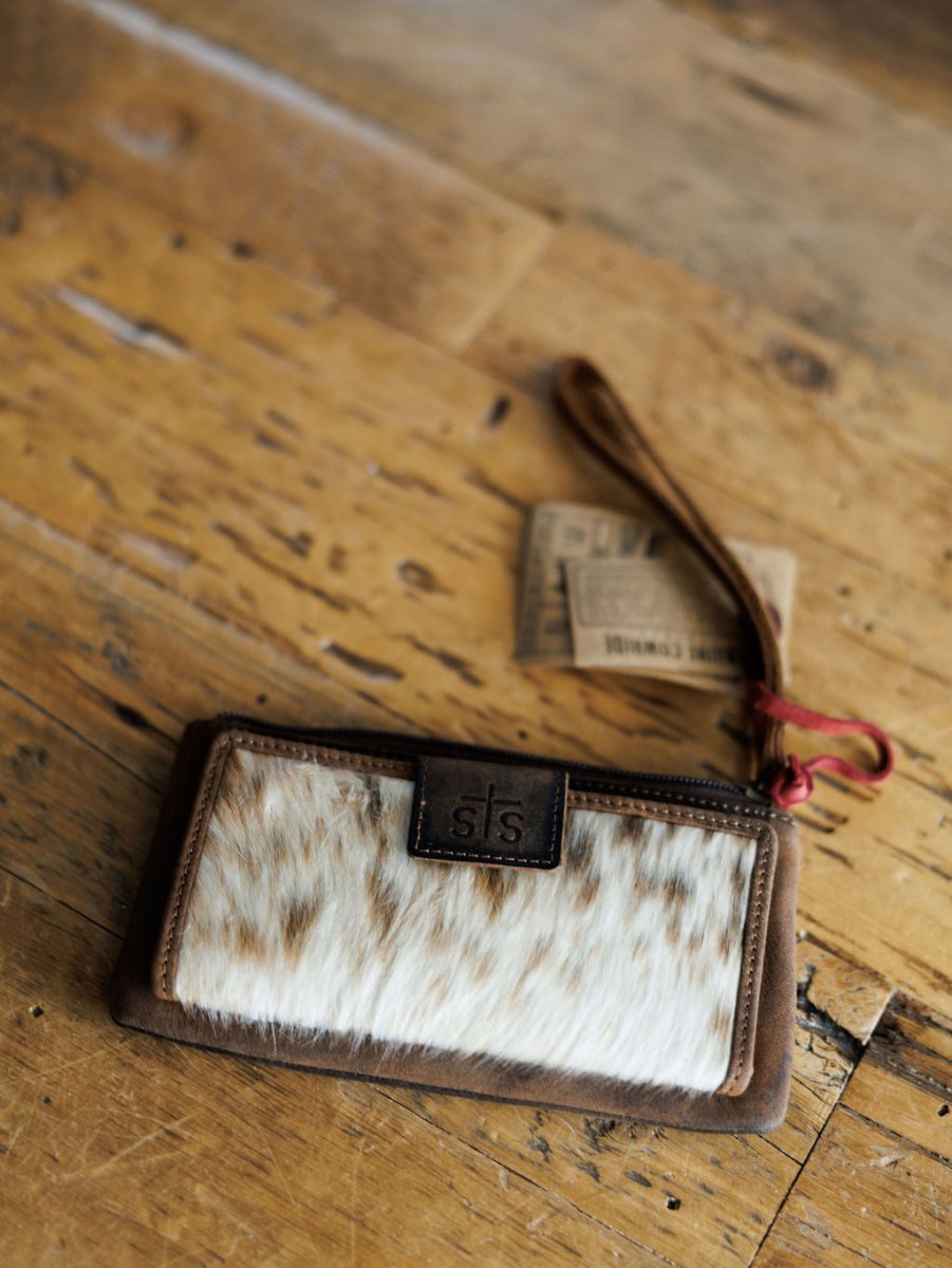 The Chisholm by Savannah Sevens western life{&}style, a cowhide wristlet wallet with leather trim, fur-like patterned front, snap closure, and a brown STS-embossed tag, rests on a wooden surface.
