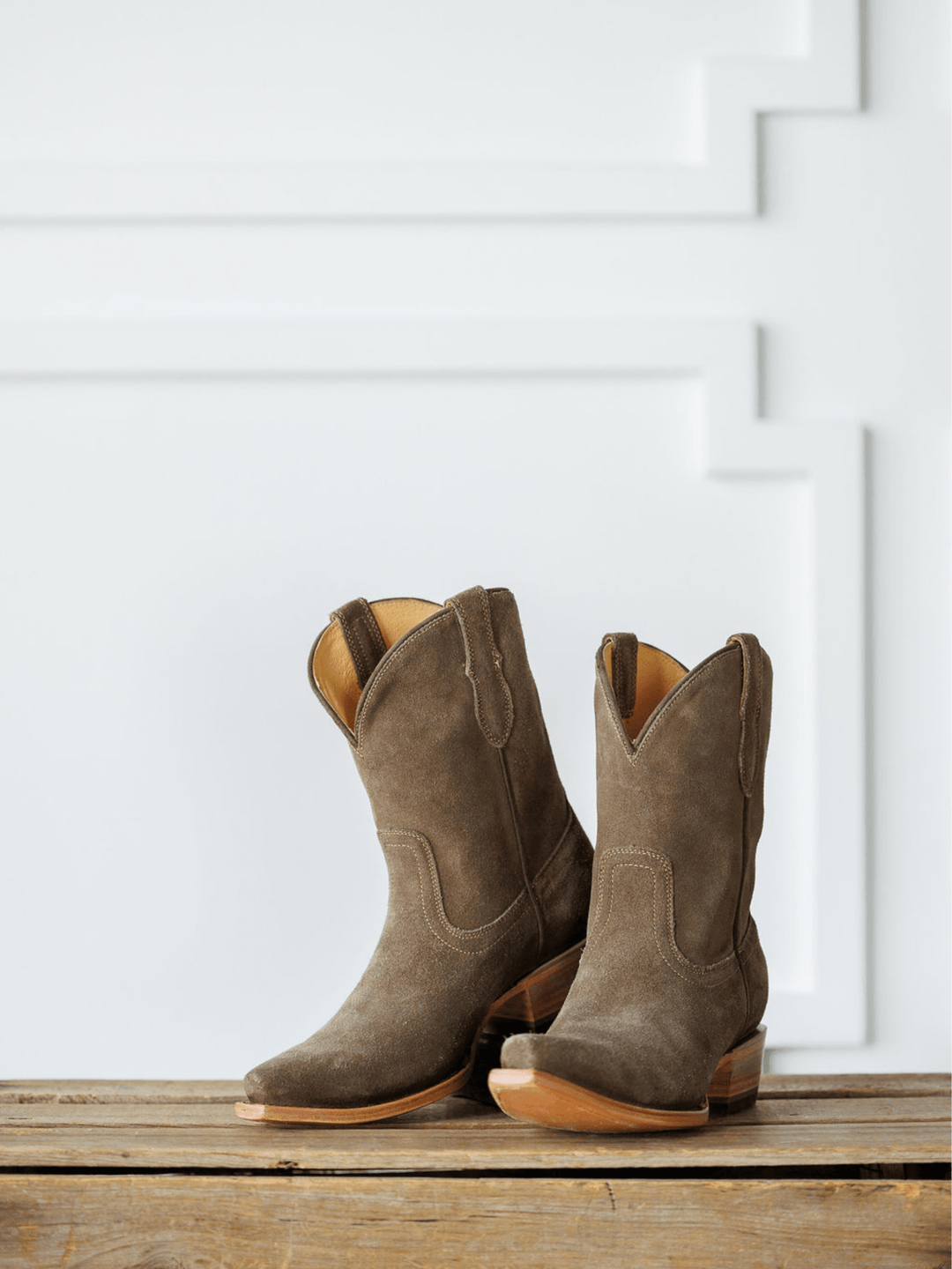 The Cordova booties by Savannah Sevens western life{&}style, crafted in brown rough out leather with wooden heels, rest on a rustic wooden surface before a white wall with geometric molding. Made by Black Star. 