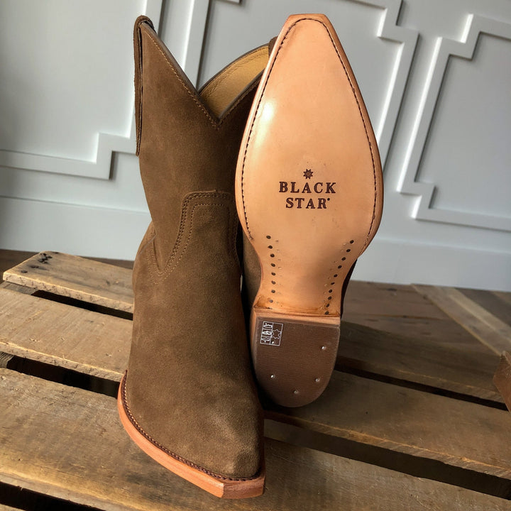 A brown suede snip toe bootie, The Cordova by Savannah Sevens western life{&}style, is propped up to display its leather sole stamped with BLACK STAR in black. It rests on wood with a decorative white wall behind it. Made by Black Star. 