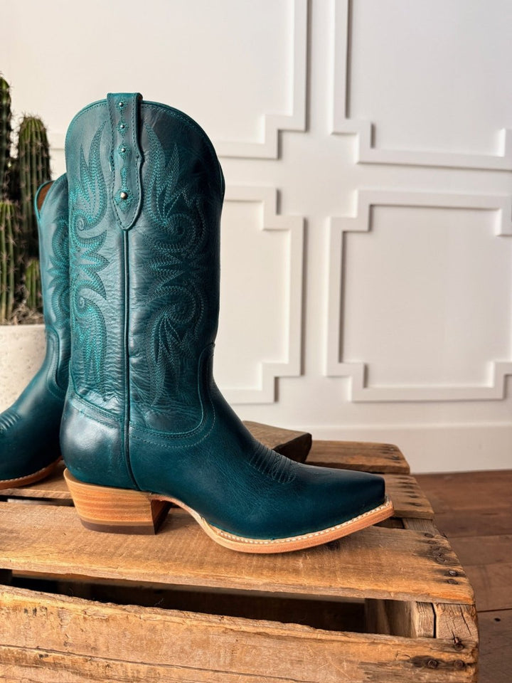A pair of The Corsicana teal leather snip toe boots by Savannah Sevens western life{&}style with decorative stitching and wooden heels are displayed on a rustic crate, set against a white paneled wall with geometric designs and a cactus. Made by Black Star. 