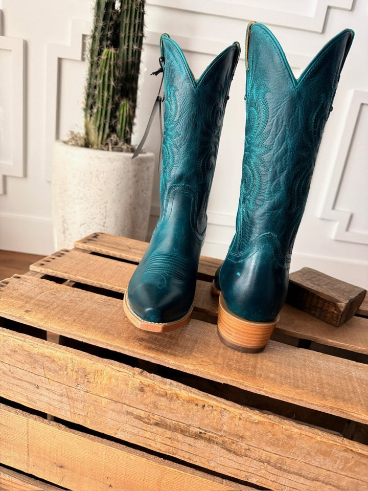 The Corsicana boots by Savannah Sevens western life{&}style, in teal leather with snip toes and decorative stitching, are displayed on a wooden pallet with a tall cactus behind them against a white paneled wall. Made by Black Star. 