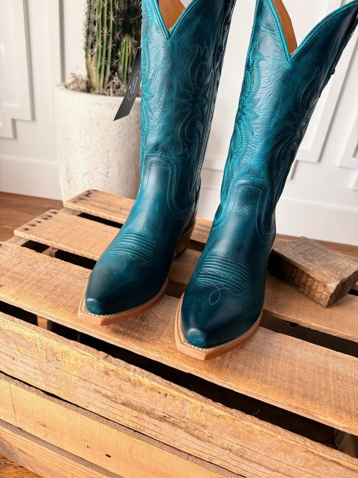 The Corsicana boots by Savannah Sevens western life{&}style, featuring teal leather and decorative stitching, stand on a wooden pallet with a tall white planter and cactus in the background. Made by Black Star. 