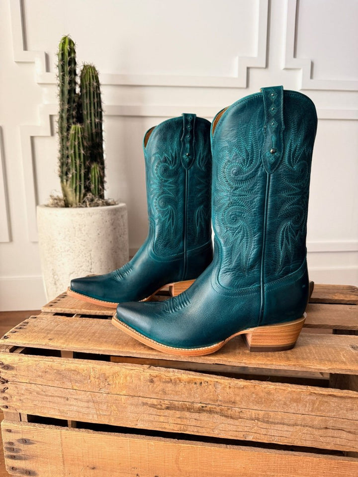 A pair of The Corsicana teal leather snip toe western boots by Savannah Sevens western life{&}style with decorative stitching rest on a wooden crate, while a tall potted cactus stands in the background against a white paneled wall.