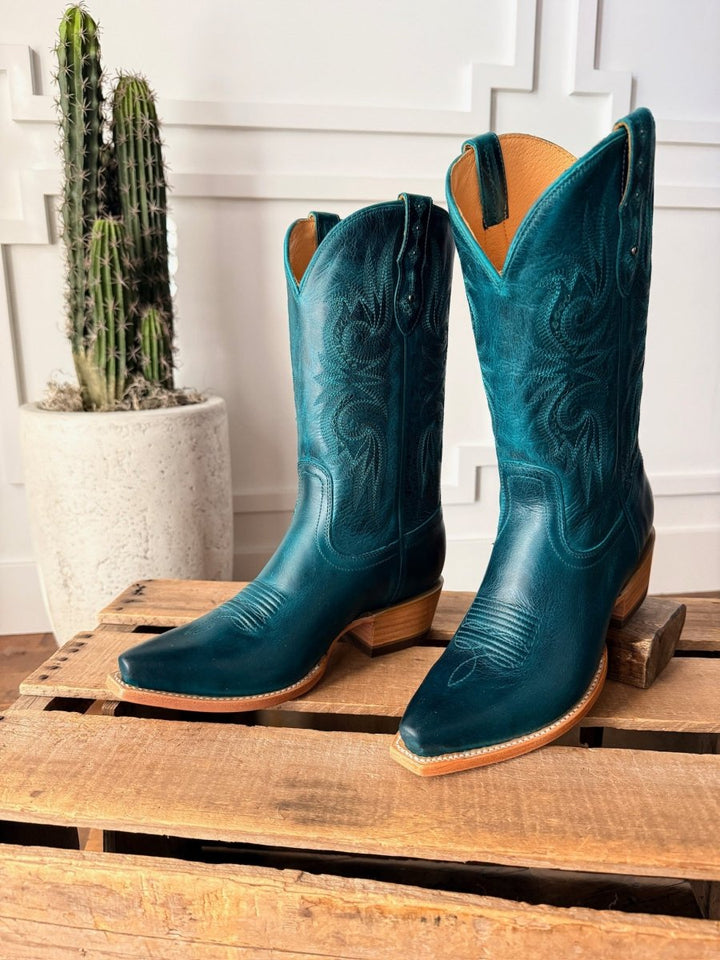 The Corsicana by Savannah Sevens western life{&}style—teal leather snip toe boots with decorative stitching—sit on a wooden pallet, with a tall cactus in a white pot against a paneled wall, embodying classic western boot style.