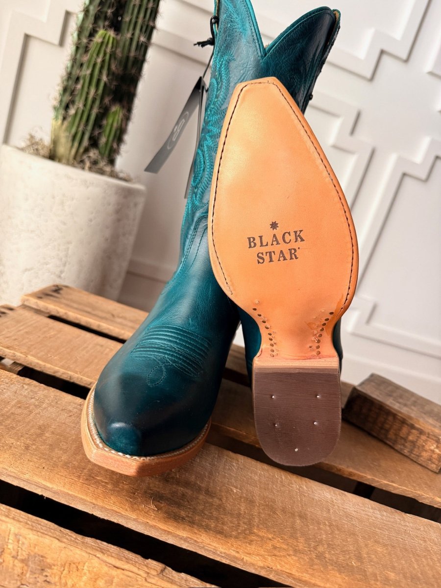 The Corsicana by Savannah Sevens western life{&}style, a teal leather snip toe western boot, stands upright on wood with its sole showing the Black Star logo. A potted cactus and a white textured wall are in the background. Made by Black Star. 