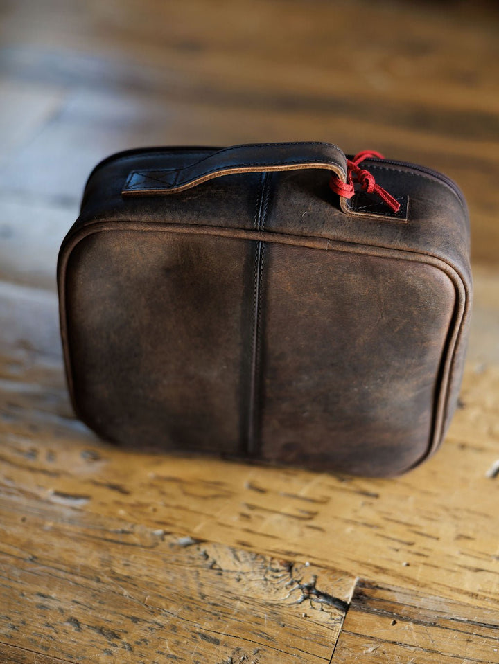 The Cowtown makeup bag by Savannah Sevens western life{&}style, featuring brown leather, a handle, and a red zipper pull, rests on a rustic wooden floor.