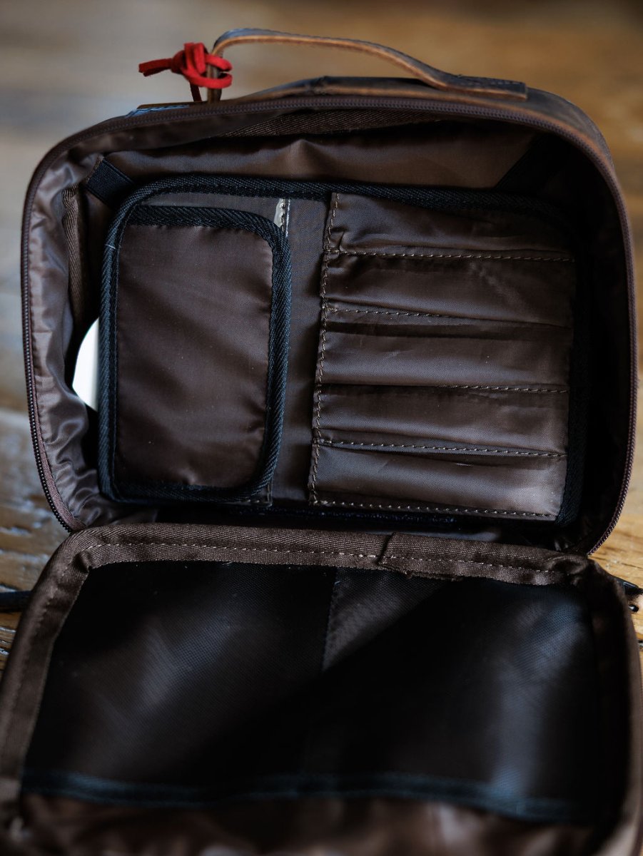 The Cowtown by Savannah Sevens western life{&}style is an open brown travel makeup case with multiple inner compartments and pockets, stitched sections for organization, and a red zipper pull visible at the top on a wooden surface.