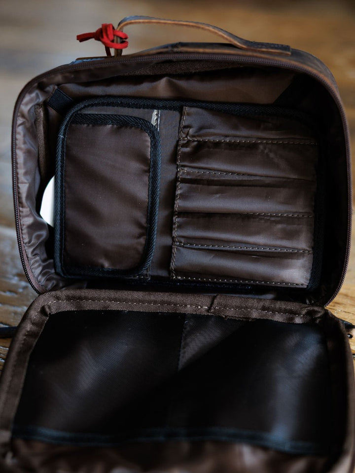 The Cowtown by Savannah Sevens western life{&}style is an open brown travel makeup case with multiple inner compartments and pockets, stitched sections for organization, and a red zipper pull visible at the top on a wooden surface.