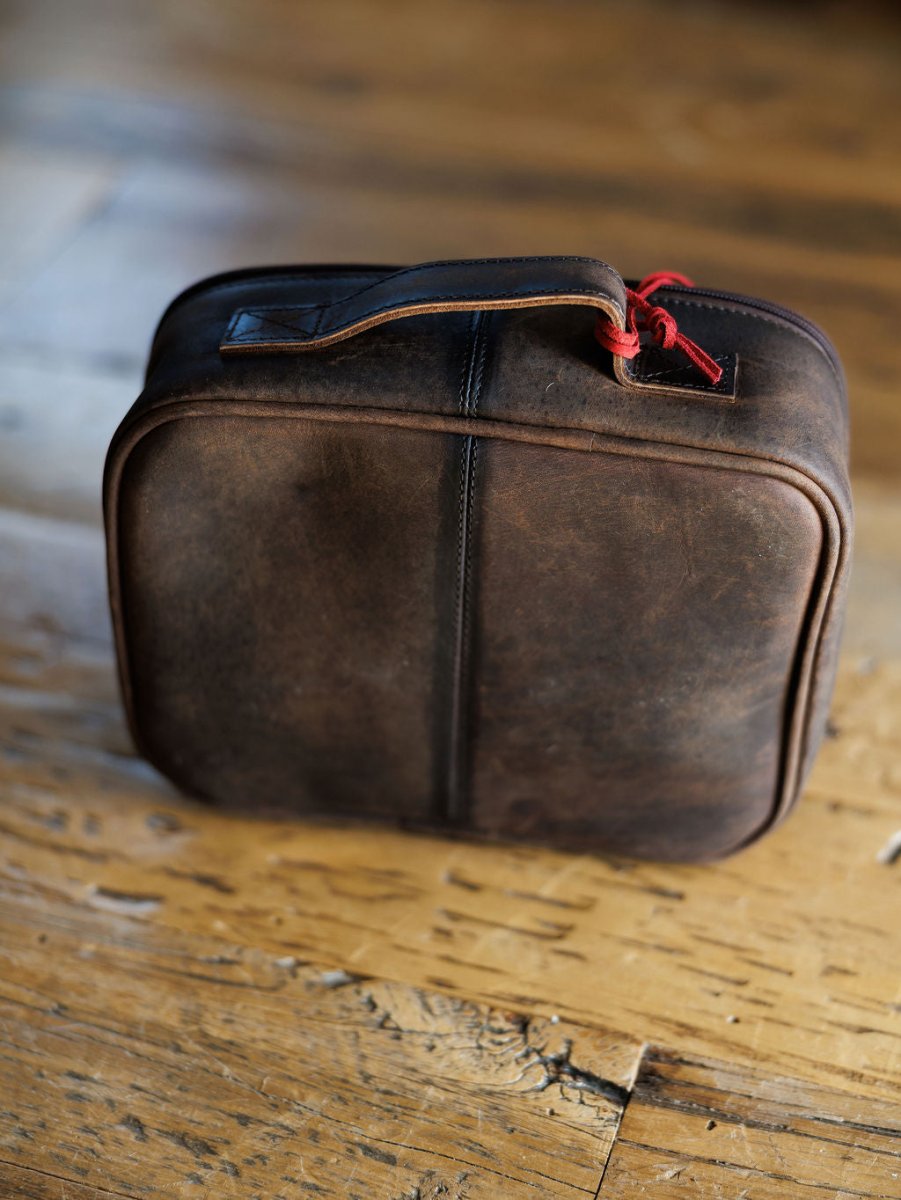 The Cowtown makeup bag by Savannah Sevens western life{&}style, featuring brown leather, a handle, and a red zipper pull, rests on a rustic wooden floor.