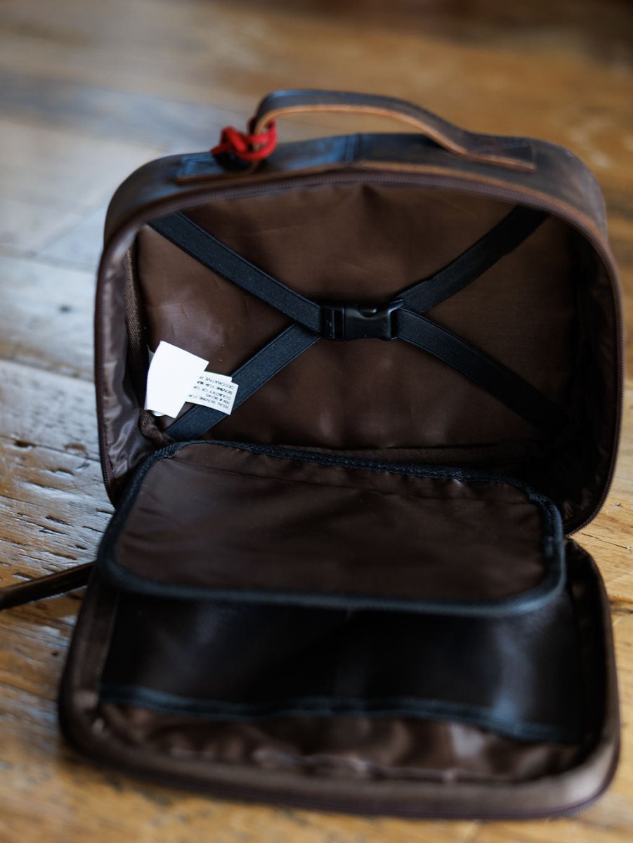 The Cowtown makeup bag by Savannah Sevens western life{&}style in brown leather with black straps and inner pocket, sits on a wooden floor. A white care label is visible inside, and the zipper is partially open.