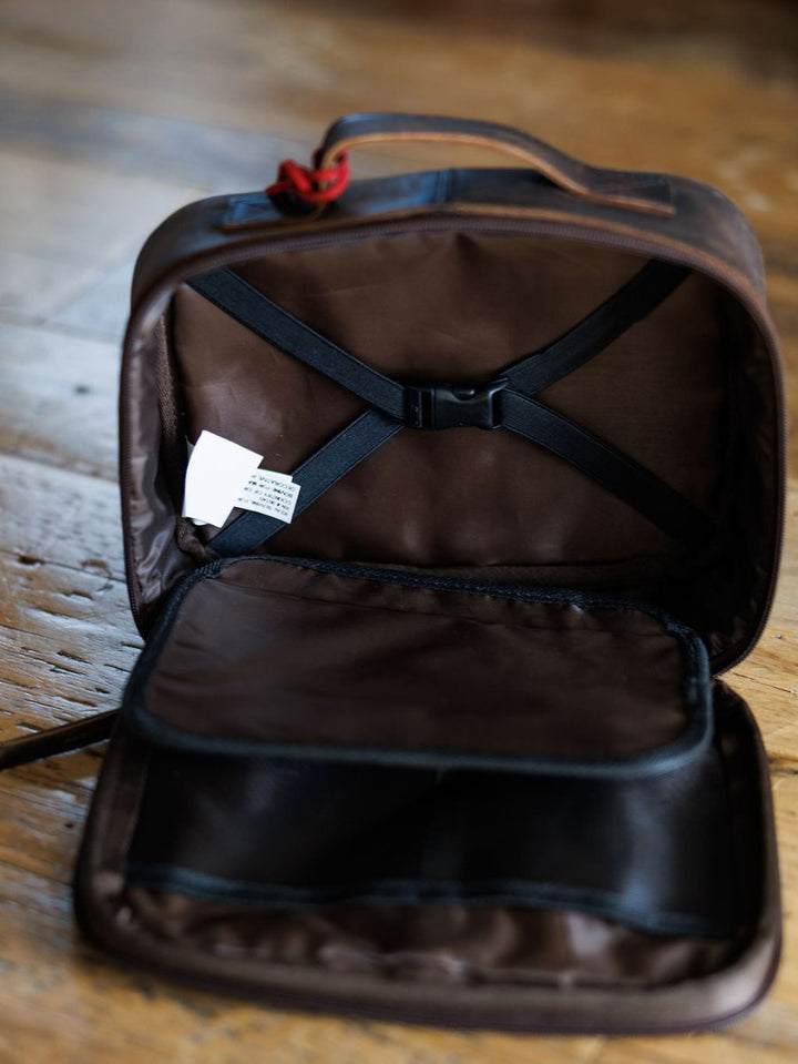 The Cowtown makeup bag by Savannah Sevens western life{&}style in brown leather with black straps and inner pocket, sits on a wooden floor. A white care label is visible inside, and the zipper is partially open.