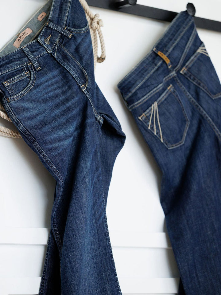 Two pairs of Ariat’s The Coy High Rise Slim Trouser Jeans in a medium dark wash hang on hooks against a white wall, one displaying the front and the other showing the back pocket.
